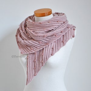 May include: A dusty rose-colored crocheted shawl with a triangular shape. The shawl features a ribbed design with small, round embellishments. The shawl is draped over a mannequin, showcasing its texture and drape. The overall aesthetic is soft and feminine.