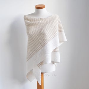 Op de afbeelding: Een witte en beige gehaakte sjaal met een driehoekige vorm. De sjaal is over een wit mannequin gelegd.