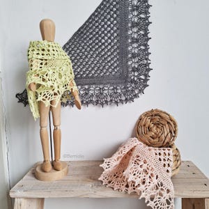 Puede incluir: Un maniquí de madera con un chal de ganchillo verde claro. Un chal de ganchillo gris cuelga de la pared. Un chal de ganchillo rosa descansa sobre una mesa de madera junto a una cesta tejida. Los chales tienen intrincados diseños calados.