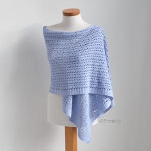 Op de afbeelding: Een lichtblauwe, open geweven gehaakte poncho, tentoongesteld op een mannequin. De poncho heeft een boothals en een vloeiende, asymmetrische zoom. Het haakpatroon creëert een delicate, luchtige textuur. Het item is zacht pastelblauw.