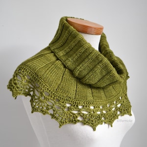 以下が含まれることがあります： オリーブグリーンのニットスヌード。リブ編みデザインと装飾的な透かし編みレースの縁取りが特徴です。マネキンに着せてあり、その質感とスタイルを示しています。肌寒い季節にぴったりの、居心地の良いアクセサリーです。