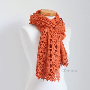 May include: A terracotta-coloured crochet scarf on a mannequin. The scarf features an open, lacy pattern and a scalloped edge. The warm, earthy tone of the scarf is highlighted by the way it is draped, showing its texture and shape.