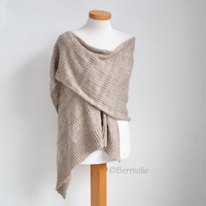 May include: A beige knitted shawl with a geometric pattern is draped over a mannequin. The shawl has a textured design and a draped neckline. The fabric appears soft and lightweight, suitable for layering. The shawl is displayed against a white background.