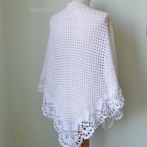 May include: A white, open-weave crochet shawl draped over a mannequin. The shawl features a delicate, scalloped lace border along the edges. The word "Berniolie" is visible in the upper left corner. The shawl is displayed against a light green background.