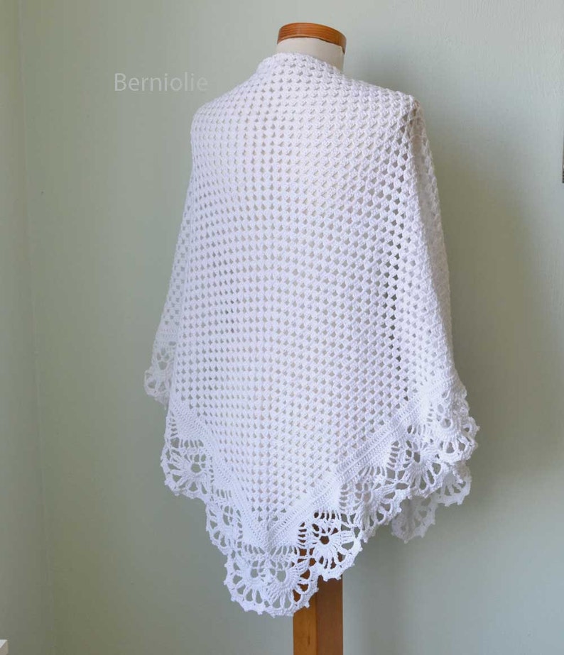 May include: A white, open-weave crochet shawl draped over a mannequin. The shawl features a delicate, scalloped lace border along the edges. The word "Berniolie" is visible in the upper left corner. The shawl is displayed against a light green background.
