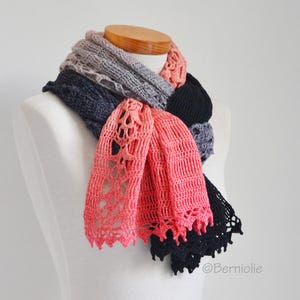 Może przedstawiać: Wielokolorowy szalik prezentowany na manekinie. Szalik posiada sekcje z szarej, granatowej, czarnej i koralowej koronki szydełkowej. Koralowa sekcja ma ażurowy wzór. Szalik jest narzucony na drewniany kształt szyi.