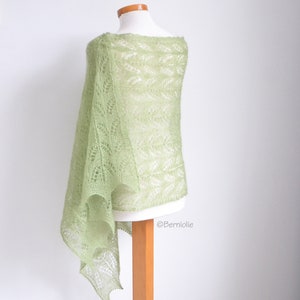 May include: A light green, open-weave shawl draped over a mannequin. The shawl has a delicate, lacy pattern and a soft, airy appearance. The fabric is a pale, spring-like green, and the shawl is displayed on a wooden stand.