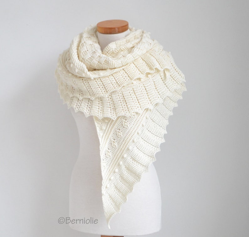 May include: An off-white knitted triangular shawl featuring a textured pattern with ribbed sections, small bobbles, and a ruffled edge. The garment is draped over a mannequin and includes the text Berniolie printed in the bottom left corner.
