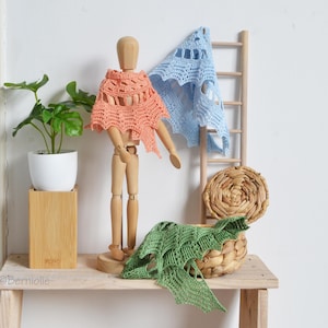 Puede incluir: Un maniquí de artista de madera exhibe un chal de ganchillo color melocotón. Un chal de ganchillo azul claro cuelga de una pequeña escalera de madera. Un chal de ganchillo verde descansa sobre una cesta de mimbre. Una planta en maceta se encuentra sobre un soporte de madera.