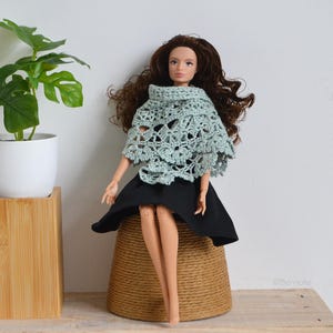 Puede incluir: Una muñeca con un chal de ganchillo verde claro y una falda negra. La muñeca tiene el pelo largo castaño y está sentada sobre una base tejida marrón. Una planta en maceta está en el fondo.