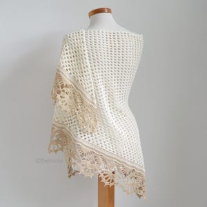 May include: A cream-colored, open-weave crochet shawl draped over a mannequin. The shawl features a delicate, scalloped edge in a contrasting tan color. The design is airy and lightweight, perfect for layering.