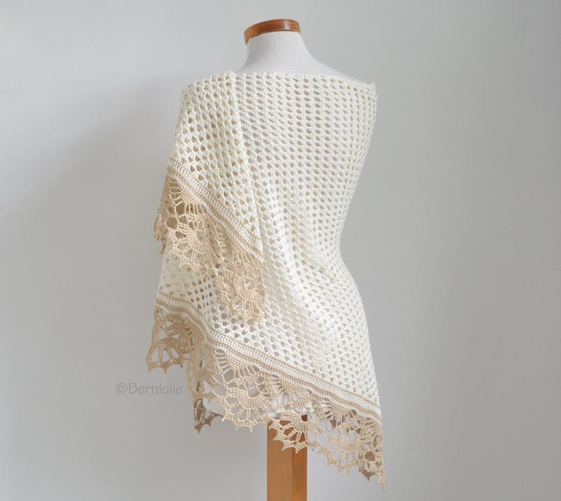 May include: A cream-colored, open-weave crochet shawl draped over a mannequin. The shawl features a delicate, scalloped edge in a contrasting tan color. The design is airy and lightweight, perfect for layering.