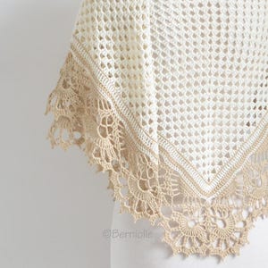 May include: A cream-colored crochet shawl with a delicate openwork design. The shawl features a contrasting tan border and a scalloped edge with intricate floral patterns. The design is visible in the image.