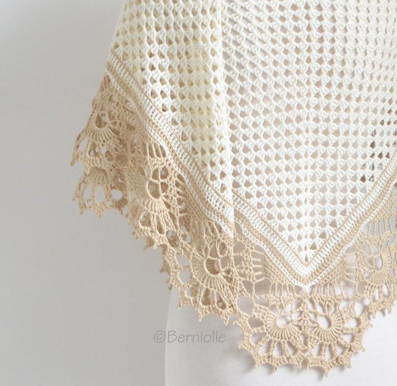 May include: A cream-colored crochet shawl with a delicate openwork design. The shawl features a contrasting tan border and a scalloped edge with intricate floral patterns. The design is visible in the image.