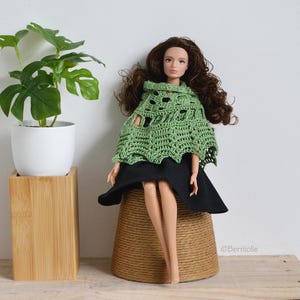 Puede incluir: Una muñeca con un poncho verde de ganchillo y una falda negra, sentada sobre una base envuelta en cuerda. La muñeca tiene el pelo largo castaño y está cerca de una planta en maceta en una maceta blanca sobre un soporte de madera.