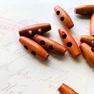 May include: A group of 10 brown wooden toggle buttons with two holes each. The buttons are oval shaped and have a smooth finish.
