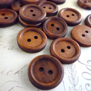 May include: A collection of brown wooden buttons with two holes each. The buttons are arranged in a random pattern on a white surface.