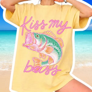 Peut inclure: T-shirt jaune avec l'inscription rose "Kiss my bass". Le t-shirt présente un graphisme d'un poisson vert, rose et orange. Le t-shirt est de style décontracté et surdimensionné.
