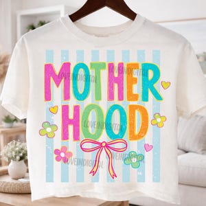 May include: Cream-colored crop top with the word "MOTHERHOOD" in colorful, playful lettering. The design includes flowers, hearts, and a bow, set against a backdrop of light blue vertical stripes. The shirt is hanging on a wooden hanger.