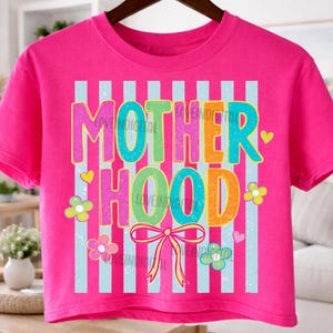 May include: A bright pink crop top with the word "MOTHERHOOD" in colorful, bubble letters. The design includes light blue vertical stripes, flowers, hearts, and a bow. The shirt is hanging on a dark brown hanger.