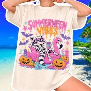 Peut inclure: T-shirt crème avec un motif coloré. Le motif représente un squelette de raton laveur portant des lunettes de soleil, se relaxant sur une bouée flamant rose, tenant une boisson. Le texte "SUMMERWEEN VIBES" est au-dessus du raton laveur. Citrouilles, bougies et chauves-souris.