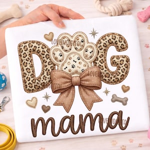 May include: White sweatshirt with the words "DOG mama" in brown embroidery. The word "DOG" features leopard print letters. A paw print design and a brown bow are centered on the sweatshirt, with small heart and bone accents.