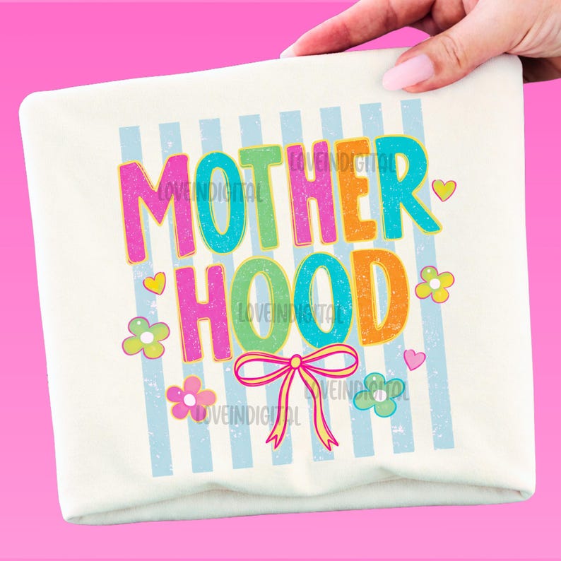 May include: A cream-colored pillow with the word "MOTHERHOOD" in colorful, bubble letters. The design includes blue vertical stripes, flowers, hearts, and a pink bow. The pillow is held by a hand.