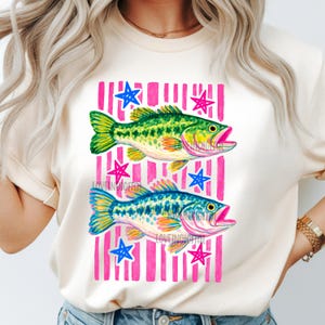 May include: Cream-colored t-shirt featuring a watercolor design of two fish, one green and one blue, set against a pink striped background with blue and pink stars. The text "LOVEINDIGITAL" is visible on the design.