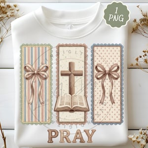 以下が含まれることがあります： 3つのフレームデザインが施された白いスウェットシャツ。デザインには、ストライプの背景にリボン、十字架と開いた本、水玉模様の背景にリボンが含まれています。「PRAY」の文字が下部に印刷されています。