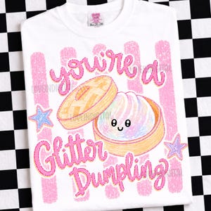 May include: White t-shirt with a pink and orange illustration of a dumpling in a bamboo steamer. The text "you're a Glitter Dumpling" is in pink script. The shirt has a crew neck and short sleeves.