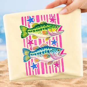 May include: A beige beach towel featuring two colorful fish illustrations. The fish are green and blue, with pink mouths and yellow accents. The design includes pink vertical stripes and blue and pink star shapes.