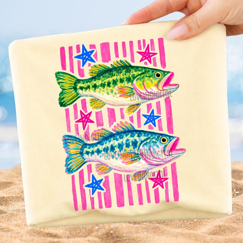 May include: A beige beach towel featuring two colorful fish illustrations. The fish are green and blue, with pink mouths and yellow accents. The design includes pink vertical stripes and blue and pink star shapes.
