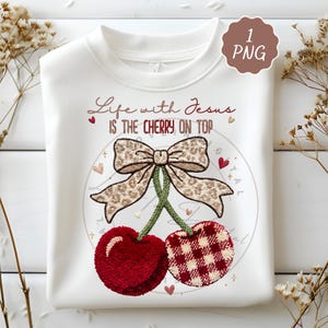 May include: White sweatshirt with a cherry design. The design features two cherries, one solid red and the other with a red and white gingham pattern, connected by green stems and a leopard print bow. Text reads "Life with Jesus is the cherry on top."