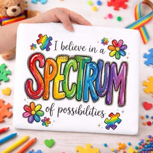 Può includere: Maglietta bianca con la scritta "I believe in a SPECTRUM of possibilities" nei colori dell'arcobaleno. Il design include pezzi di puzzle e fiori colorati. La maglietta è tenuta da una mano.