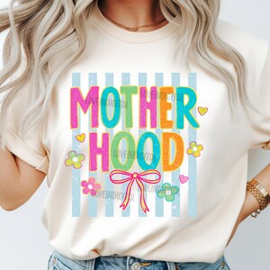 May include: Cream-colored t-shirt with the word "MOTHERHOOD" in colorful, outlined letters. The design includes flowers, hearts, and a bow, set against a backdrop of light blue stripes. The shirt is a casual wear item.