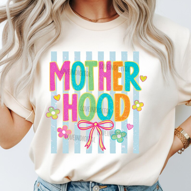 May include: Cream-colored t-shirt with the word "MOTHERHOOD" in colorful, outlined letters. The design includes flowers, hearts, and a bow, set against a backdrop of light blue stripes. The shirt is a casual wear item.