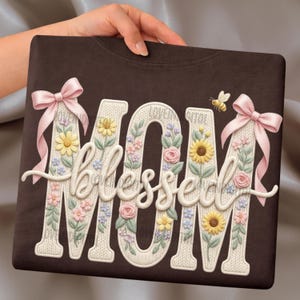 Könnte beinhalten: Ein braunes Sweatshirt mit den Worten "MOM blessed MOM" in einem floralen Design. Die Buchstaben sind weiß mit einer Strickstruktur und mit Blumen, Schleifen und einer Biene verziert.