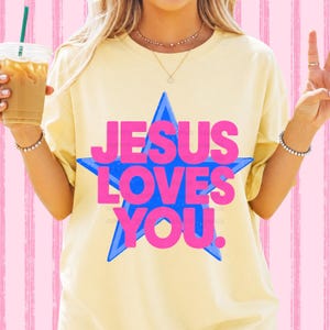 May include: A light yellow t-shirt with the text "JESUS LOVES YOU" in pink letters inside a blue star. The shirt is a casual style, suitable for everyday wear.