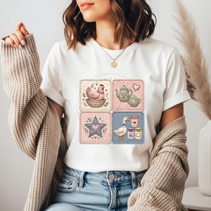 Puede incluir: Camiseta blanca con cuatro parches cuadrados con ilustraciones de tem&aacute;tica campestre. Los parches incluyen una gallina, teteras, una estrella y un ganso con tarros de mermelada. El dise&ntilde;o tiene una est&eacute;tica vintage y r&uacute;stica.