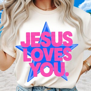 May include: Cream-colored t-shirt featuring the text "JESUS LOVES YOU" in bright pink letters, set against a blue star. The shirt has short sleeves and a relaxed fit, suitable for casual wear.