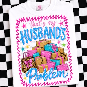 May include: White t-shirt with a graphic that reads "That's my Husband's Problem" in blue and pink. The graphic includes illustrations of gift boxes, shopping bags, and stars. The shirt has a crew neck and short sleeves.