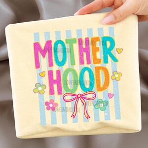 May include: A light yellow rectangular item with the word "MOTHERHOOD" in colorful, bubble letters. The design includes blue vertical stripes, flowers, hearts, and a pink bow.