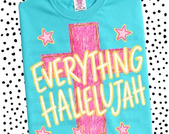 May include: A turquoise t-shirt with a pink cross design and the words "EVERYTHING HALLELUJAH" in yellow. The design includes pink stars. The shirt has a tag in the upper left corner.
