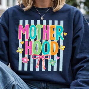 May include: Navy blue sweatshirt with the word "MOTHERHOOD" in colorful, bubble letters. The design includes vertical light blue stripes, flowers, hearts, and a bow. The text "LOVE DIGITAL" is also visible.