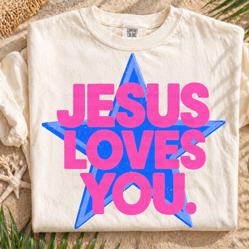 May include: Cream-colored t-shirt with the text "JESUS LOVES YOU" in bright pink letters, set against a blue star design. The shirt is made of a soft material and has a classic crew neck.