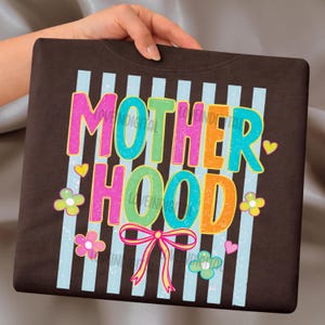 May include: A dark brown sweatshirt with the word "MOTHERHOOD" in colorful, outlined letters. The design includes light blue vertical stripes, flowers, hearts, and a pink bow. The text "LOVE DIGITAL" is also visible.