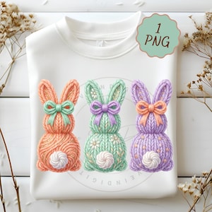 May include: White sweatshirt featuring three knitted bunny designs in orange, green, and purple. Each bunny has a bow and a fluffy white tail. The image includes a green and white "1 PNG" graphic.