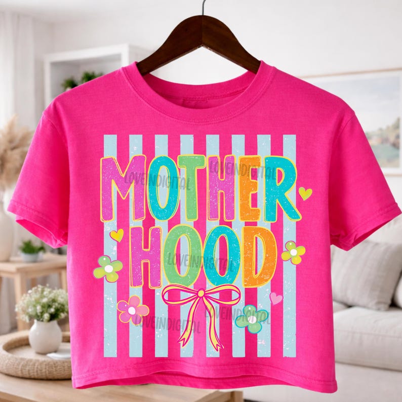 May include: A bright pink crop top with the word "MOTHERHOOD" in colorful, bubble letters. The design includes light blue vertical stripes, flowers, hearts, and a bow. The shirt is hanging on a dark brown hanger.