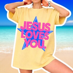 May include: Yellow t-shirt with the text "JESUS LOVES YOU" in pink letters inside a blue star. The shirt is a light yellow color and has short sleeves. The background is a beach scene with blue water and sand.