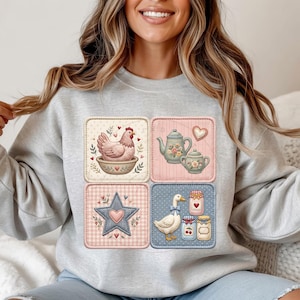 Puede incluir: Una sudadera gris claro con cuatro parches cuadrados con ilustraciones de tem&aacute;tica campestre. Los parches representan una gallina en un cuenco, teteras, una estrella y un ganso con tarros de mermelada. Las ilustraciones son en colores pastel suaves.